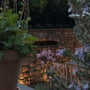 Light By Juul Cosmos chaussesten med varmt, indirekte lys, der oplyser en stenmur og planter i en have om aftenen.