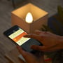 En person holder en smartphone med Philips Hue appen åben, der viser forskellige rum og lysindstillinger, rettet mod en tændt kertepære.