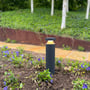 Light By Juul Platin slim pullert lyser mellem blomster og planter i et udendørs bed.
