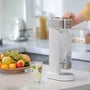En person bruger den hvide Philips sodavandsmaskine til at lave danskvand ved et køkkenbord med frugtskål og glas.