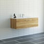 Dansani Mido møbelpakke på 121 cm med varm eg-finish og en skuffe, monteret på en hvid væg i et moderne badeværelsesmiljø med fliser.