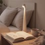 Den hvide LED-bordlampe lyser ved en seng, hvor en åben bog og en kop står på bordet.