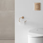 Grohe Essentials papirholder i kobberfarve monteret på en hvid væg ved siden af en toiletkumme og et kontrolpanel.