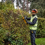 Mand i sikkerhedsudstyr bruger DeWalt 18V XR hækkeklipper med 55 cm blad på en stor busk i en have.
